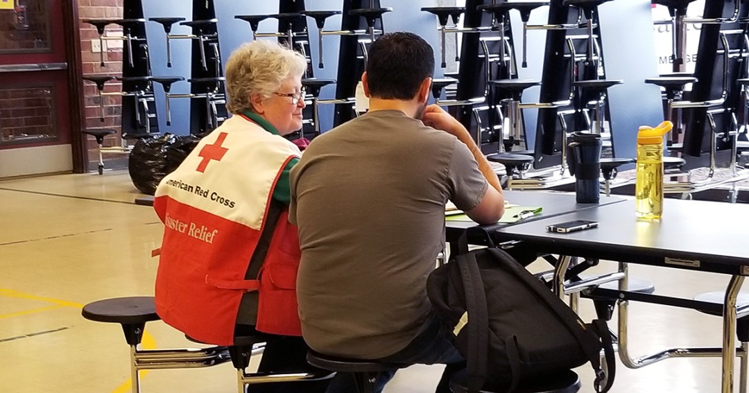 Red Cross volunteer sits next to a man at a table in a shelter.