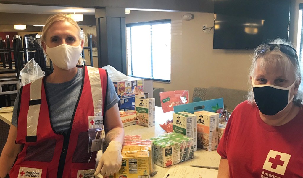 Close up photo of two red cross volunteers wearing masks standing in a kitchen in front of a counter that has pantry food items on it.